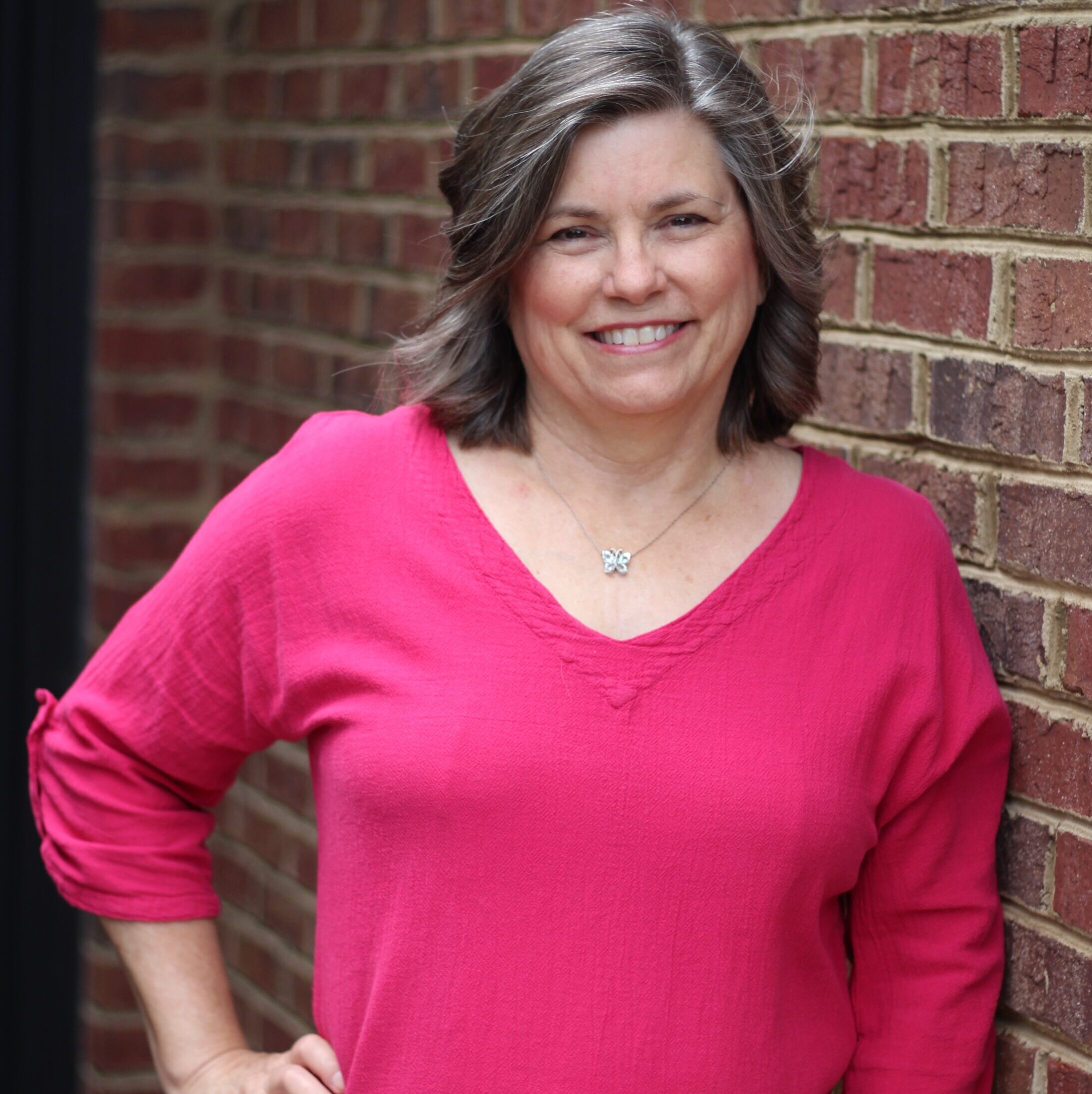 Woman in bright pink v-neck sweater smiling at camera, standing against a red brick wall with a silver pendant necklace.
