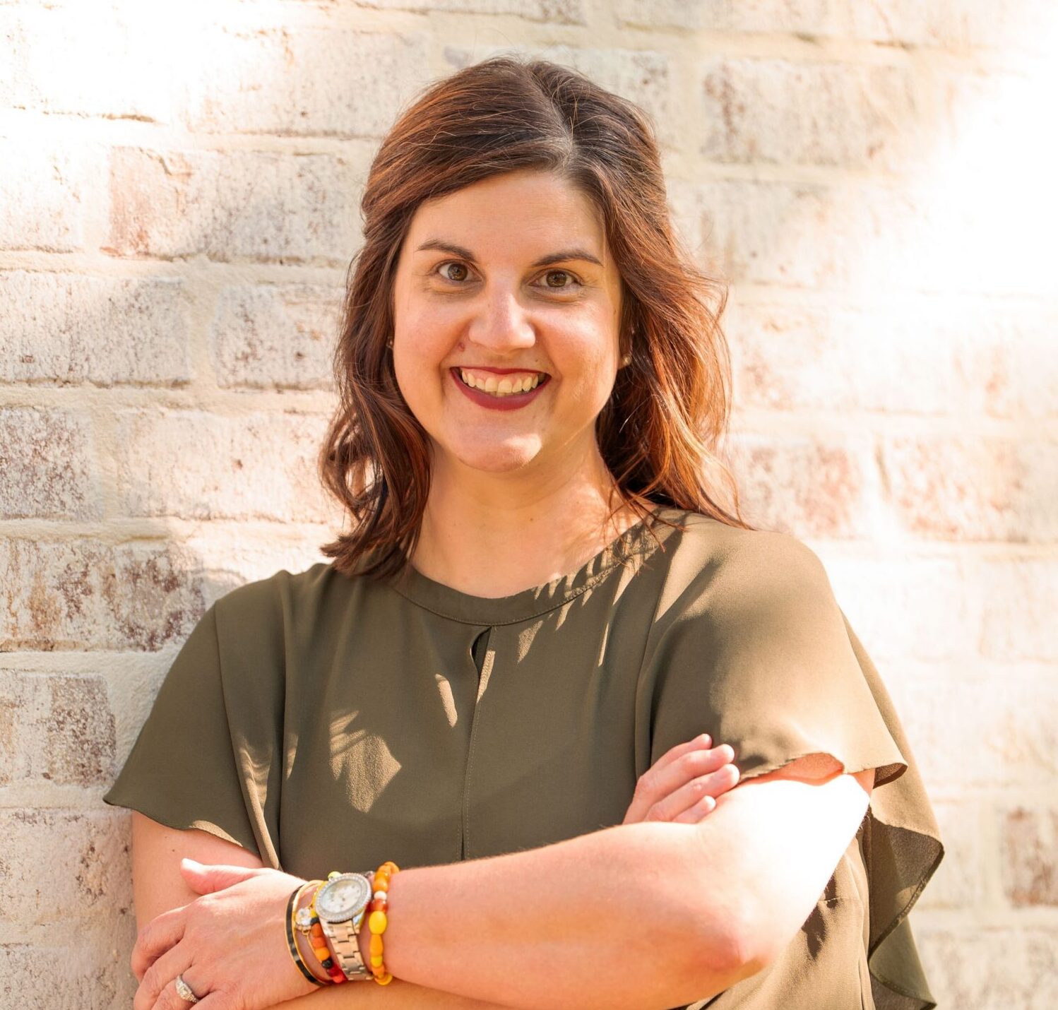 Woman with brown hair, arms crossed, wearing olive green top and colorful bracelets, standing against a white brick wall.